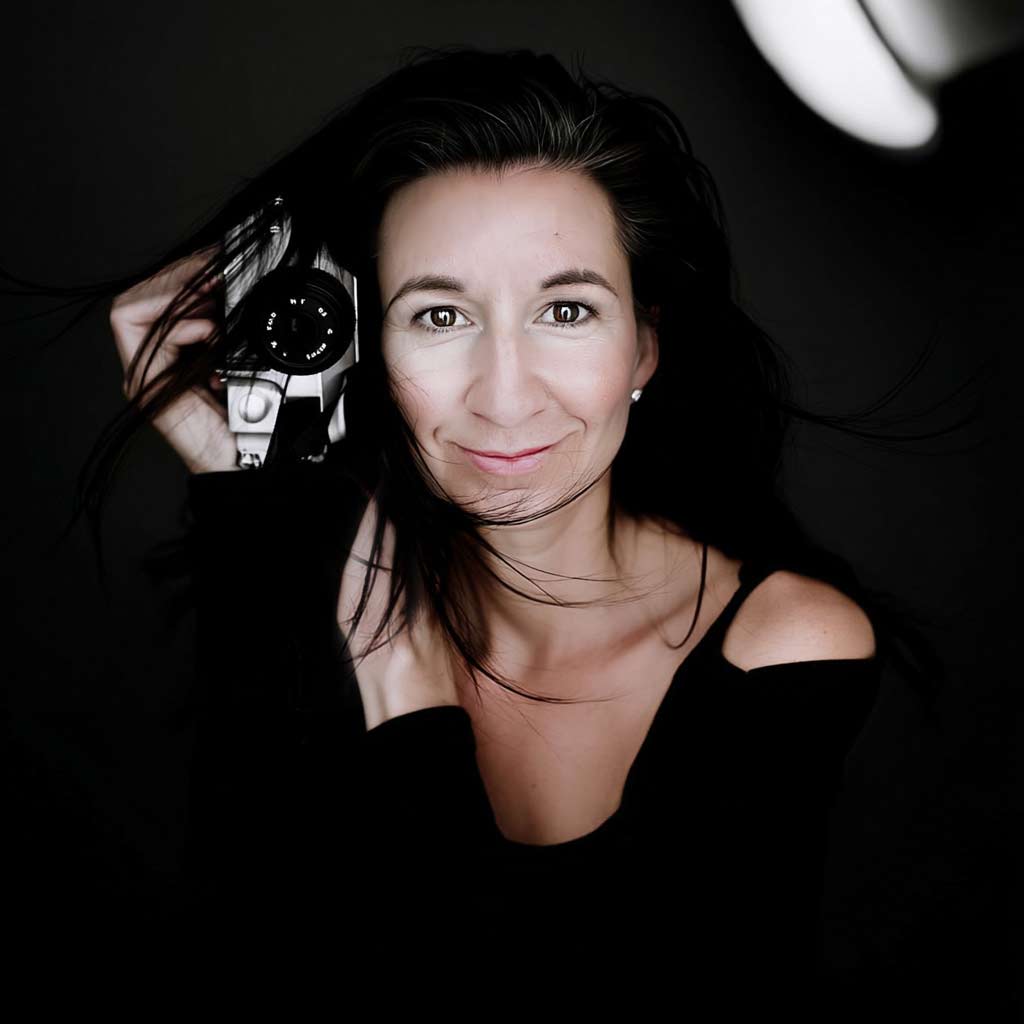 Portrait-of-Shelly-AI-Design-Expert porträt einer Frau in Weiß vor schwarzem Hintergrund, hält eine Kamera – Einblick in Shellys KI Design-Welt<br />